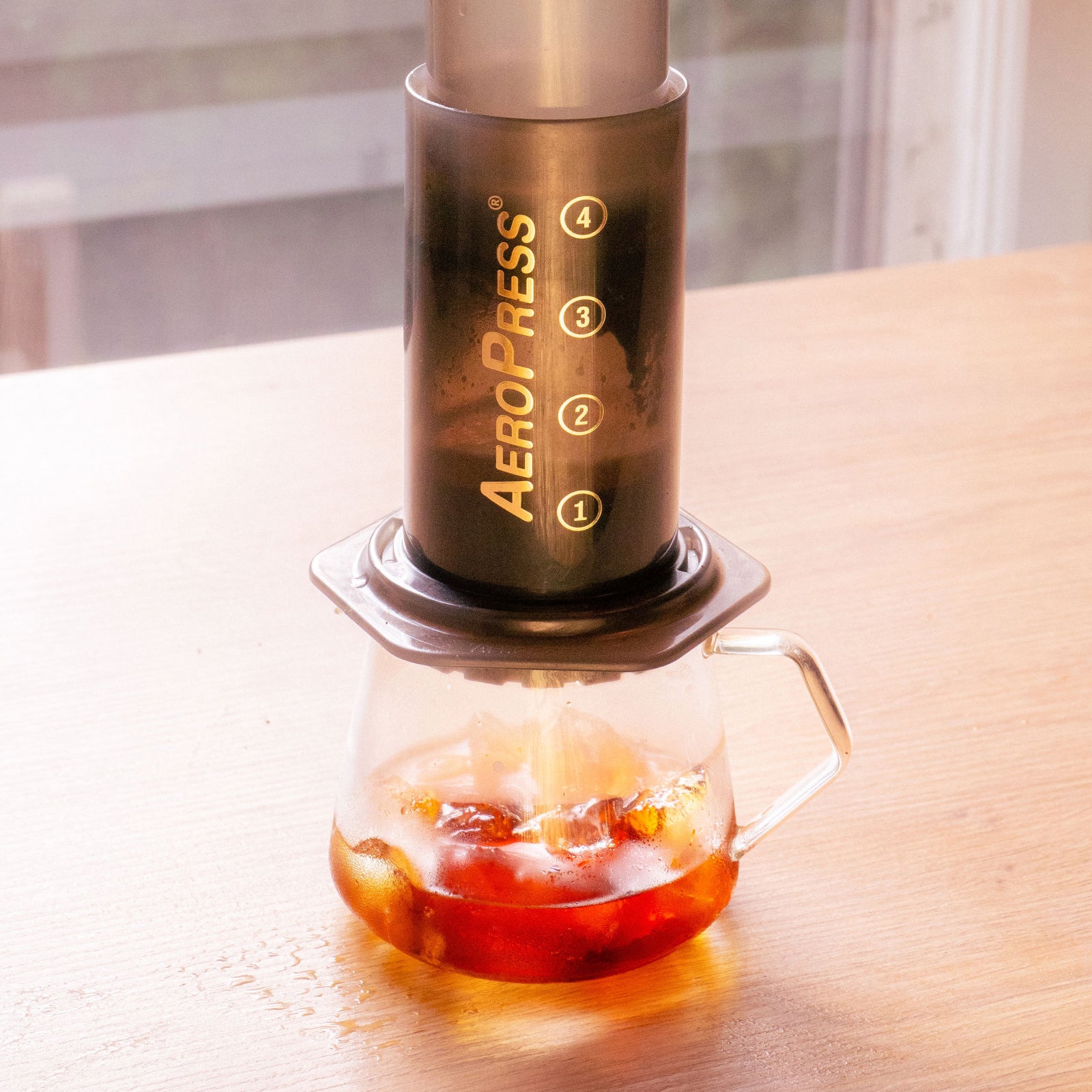 Original AeroPress coffee maker with vertical measurement marks on a dark body sits atop a glass carafe with brewed coffee
