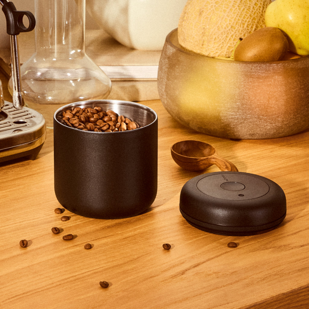 Open Atmos vacuum canister filled with roasted coffee beans on a wooden countertop with the lid beside it