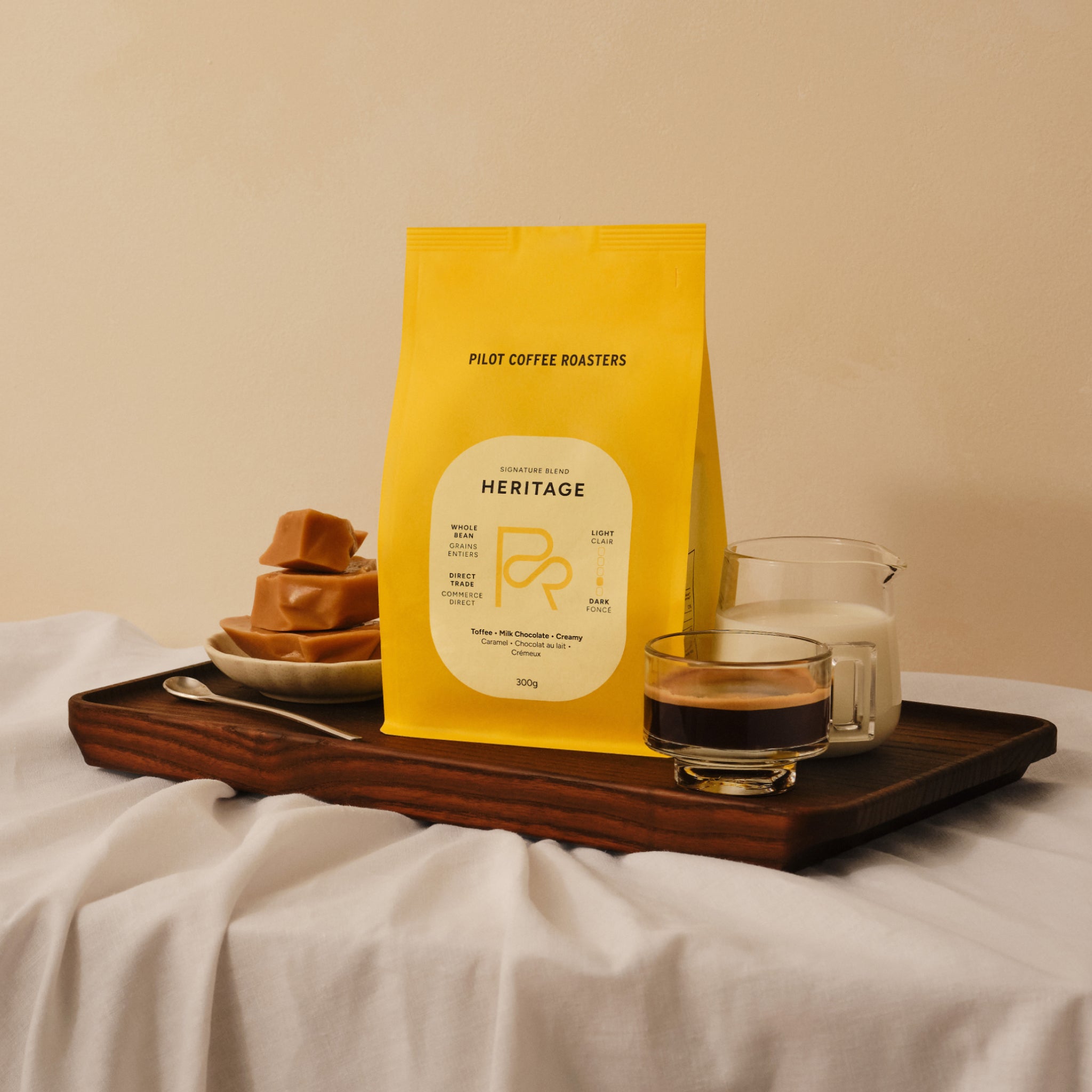 Yellow Pilot Coffee Roasters Heritage coffee bag on a dark wood tray next to a small plate of caramel candies, a shot of espresso and a small pitcher of milk