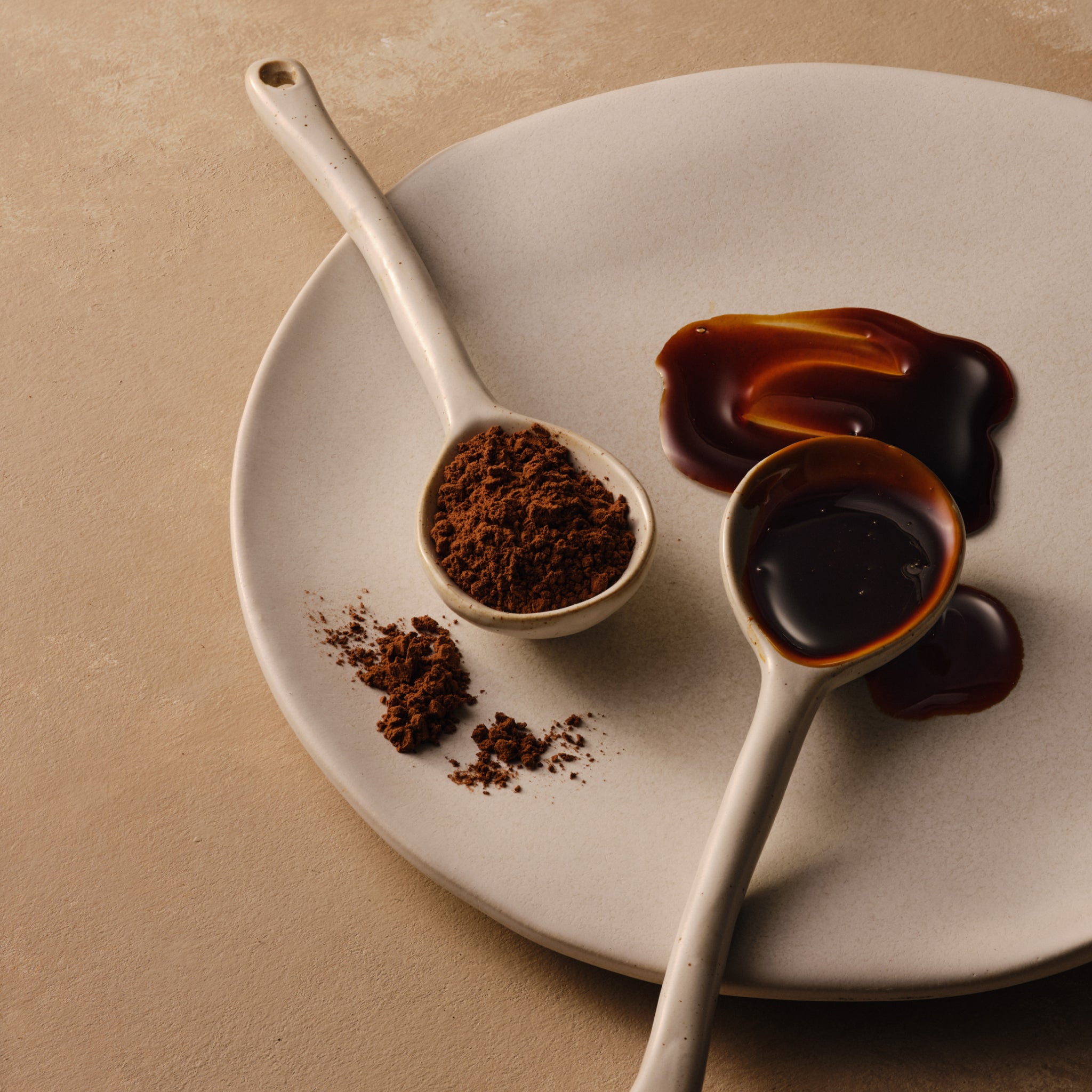 Two ceramic spoons hold cocoa powder and chocolate sauce on a white plate against a beige backdrop.
