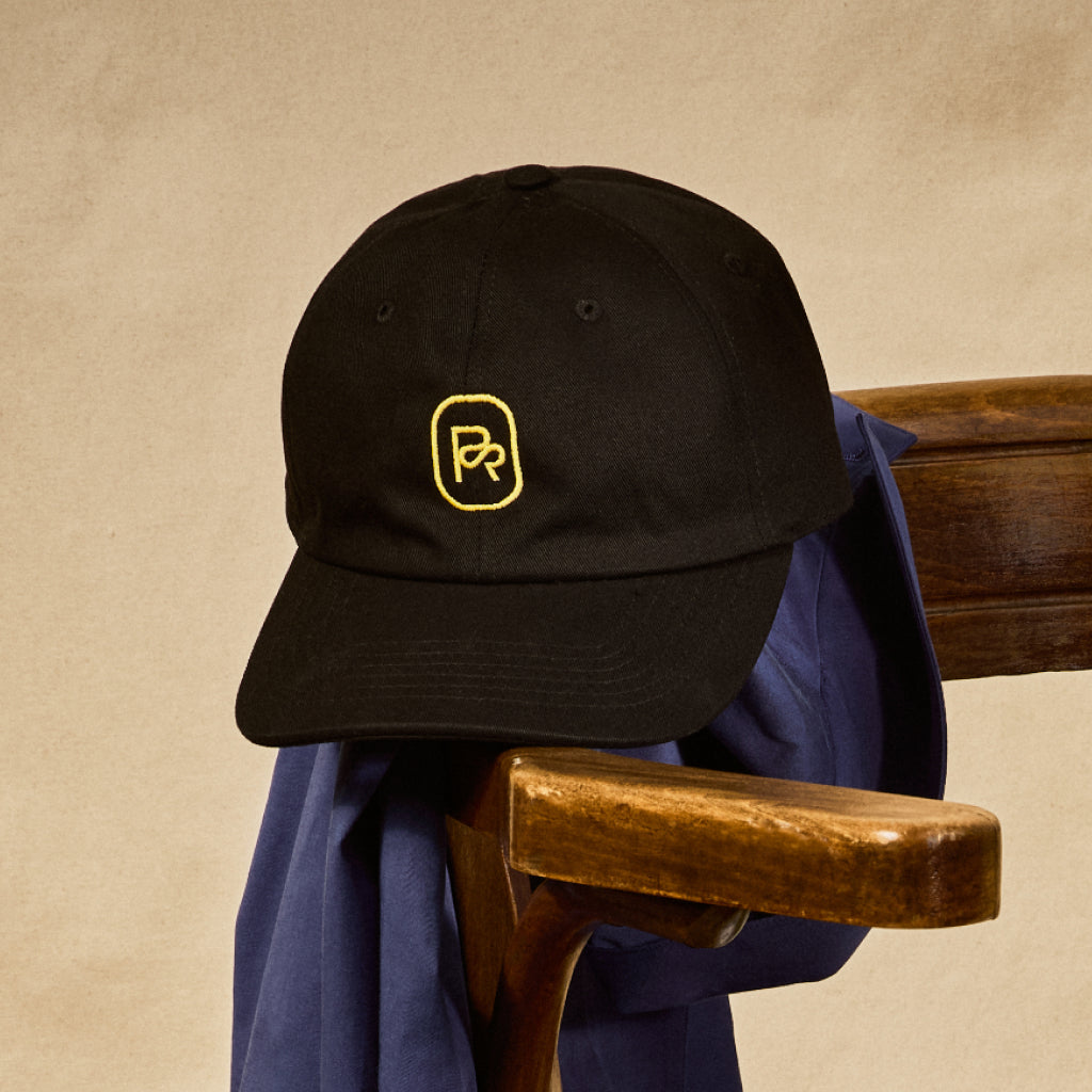 Black six panel Pilot Coffee cap with gold embroidered PCR logo on the front, resting on a wooden chair with a blue jacket draped over it