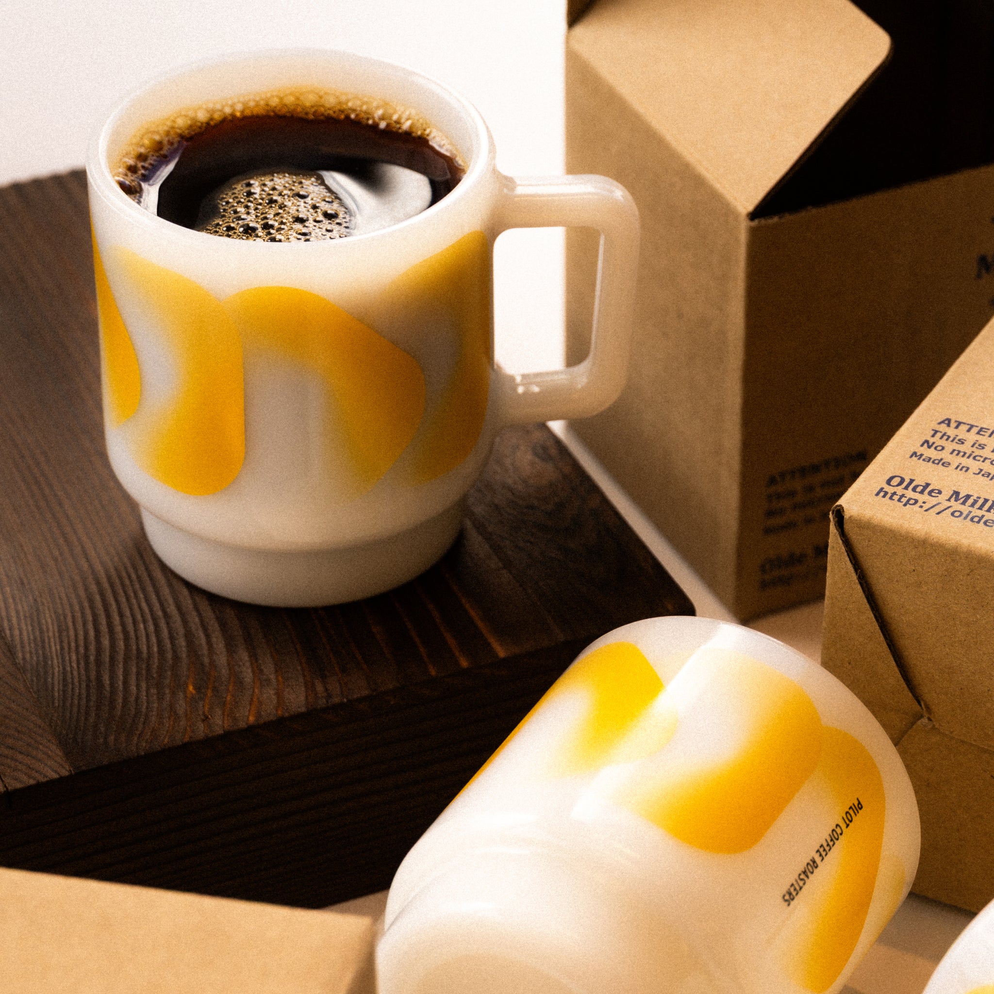 PCR branded Olde Milk-Glass mug with yellow oval pattern, filled with coffee on a wooden surface beside the product package