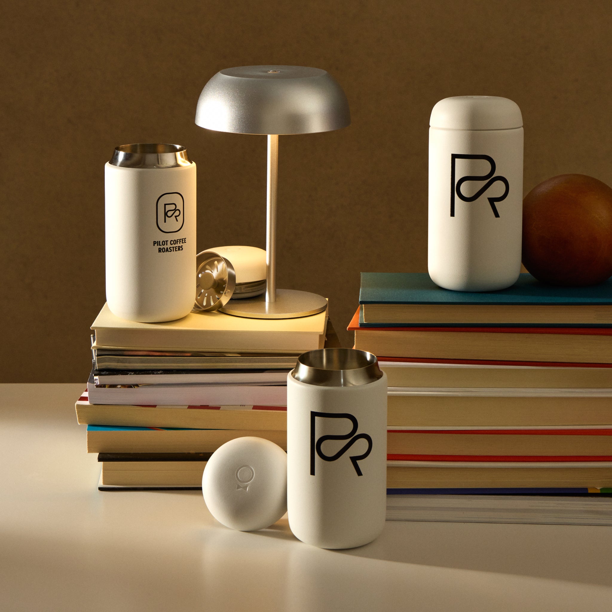 Three Pilot Coffee Roasters white travel mugs with black logo on a desk beside a lamp and stacks of books