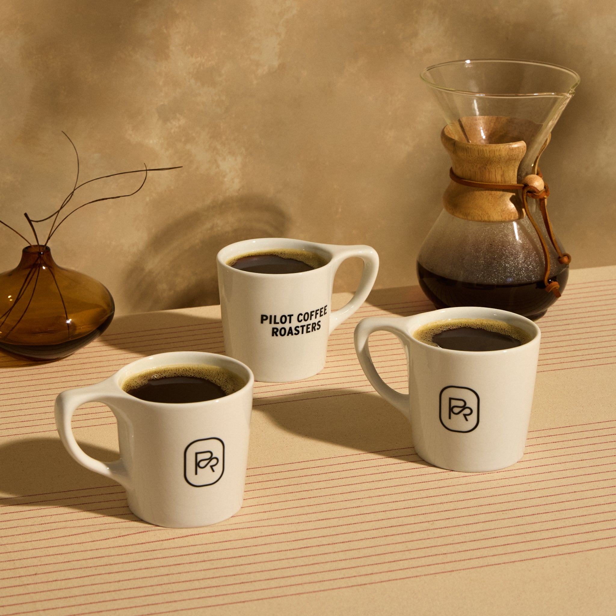 Three White 12 oz Pilot Coffee mugs with logo beside a Chemex full of brewed coffee.