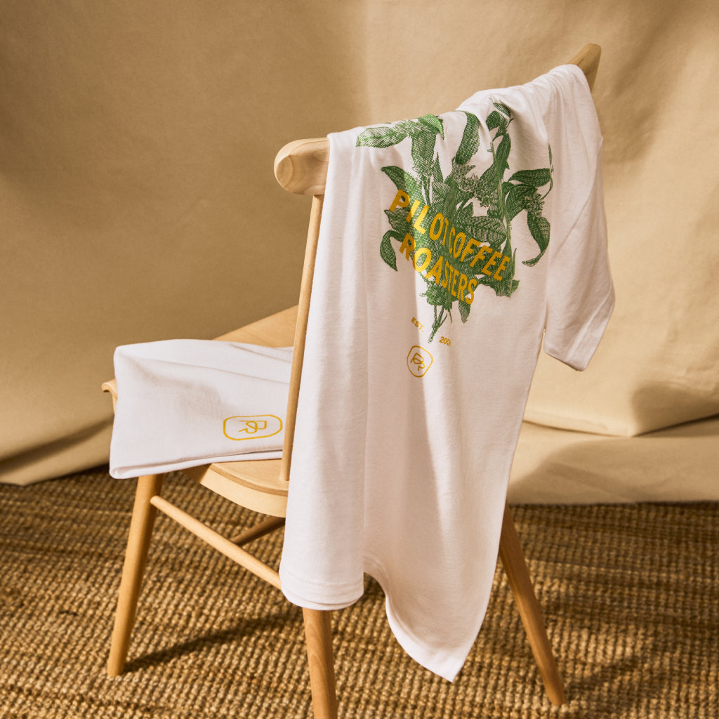 White Arabica T-shirt with green coffee plant graphic and yellow PCR logo draped over a wooden chair against a beige backdrop.