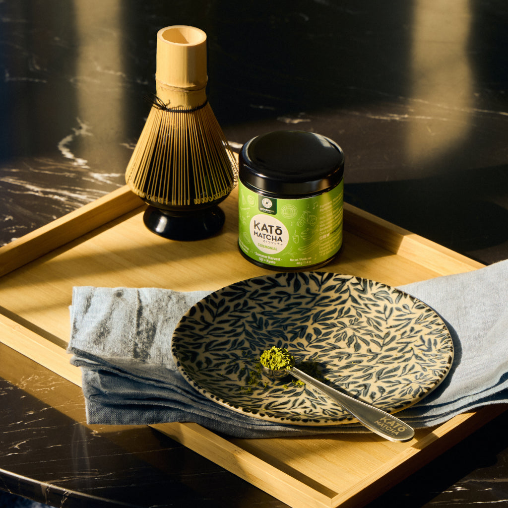 Green labeled Kato matcha tin with a bamboo whisk on a whisk stand, and matcha scoop on a wooden serving tray.