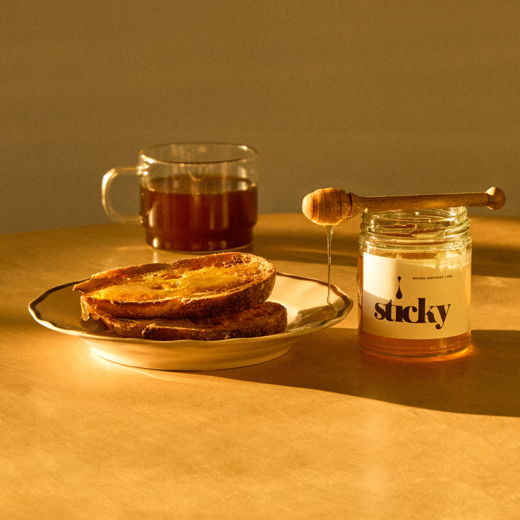 Sticky Honey jar with a wooden dipper beside toasted bread and a mug of coffee on a warm tabletop.