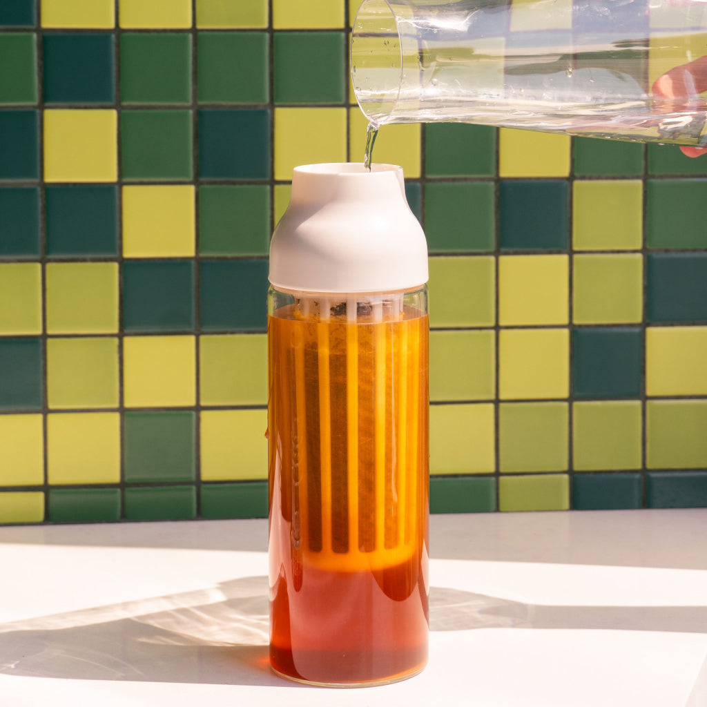 Tall glass carafe with white lid and built in filter, filled with brew, resting on a white countertop against a green and yellow tiled wall