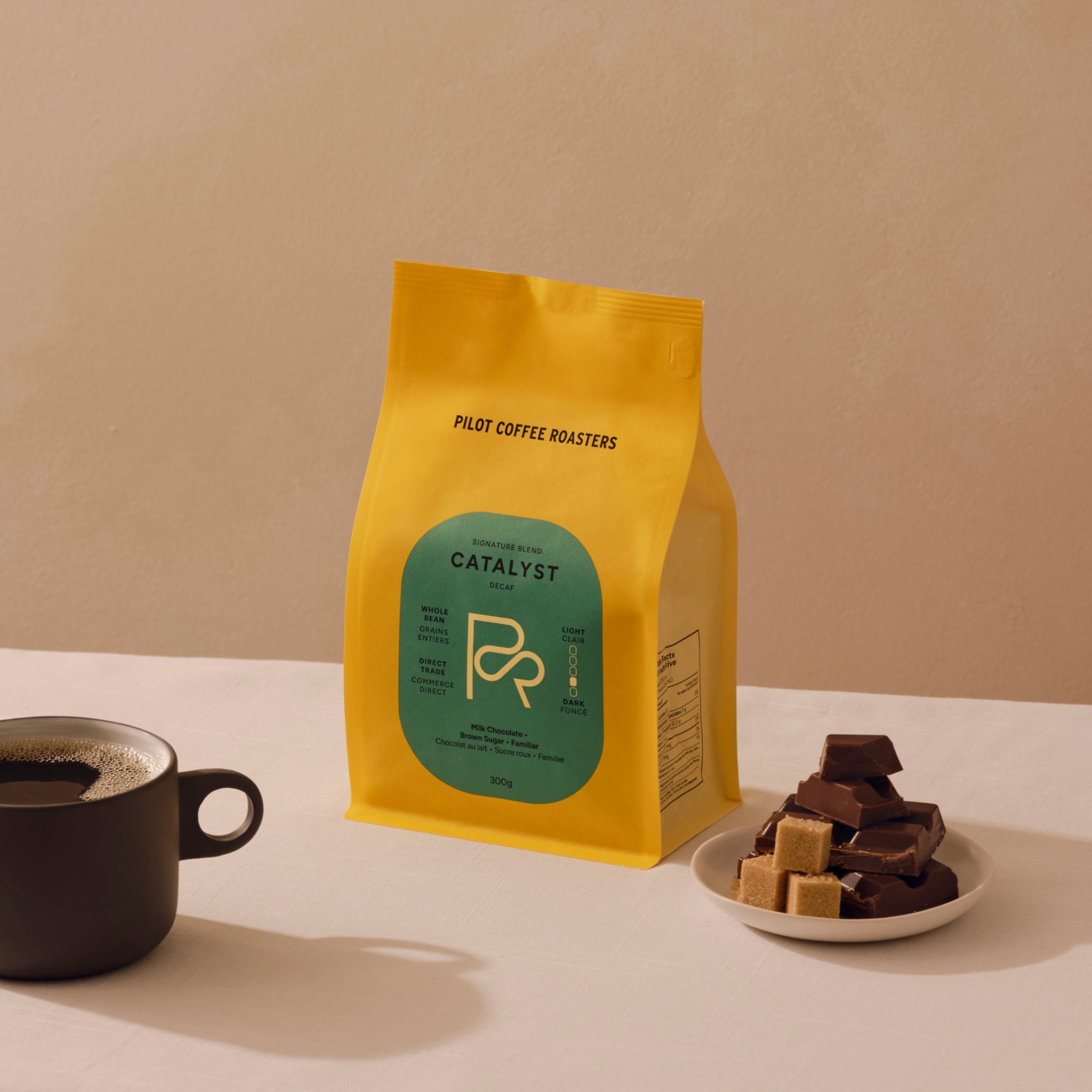 Bright yellow Pilot Coffee Roasters Catalyst Decaf bag with a green label, next to coffee mug and a saucer with brown sugar cubes.