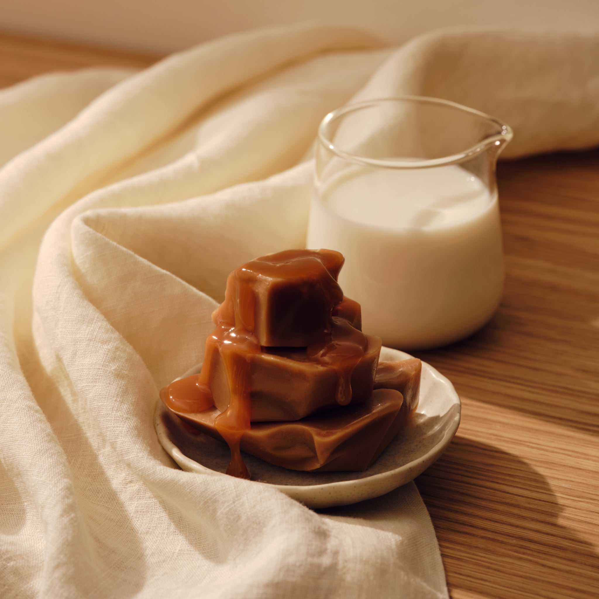 Small glass pitcher of milk behind a plate of caramel candies, surrounded by a cream cloth