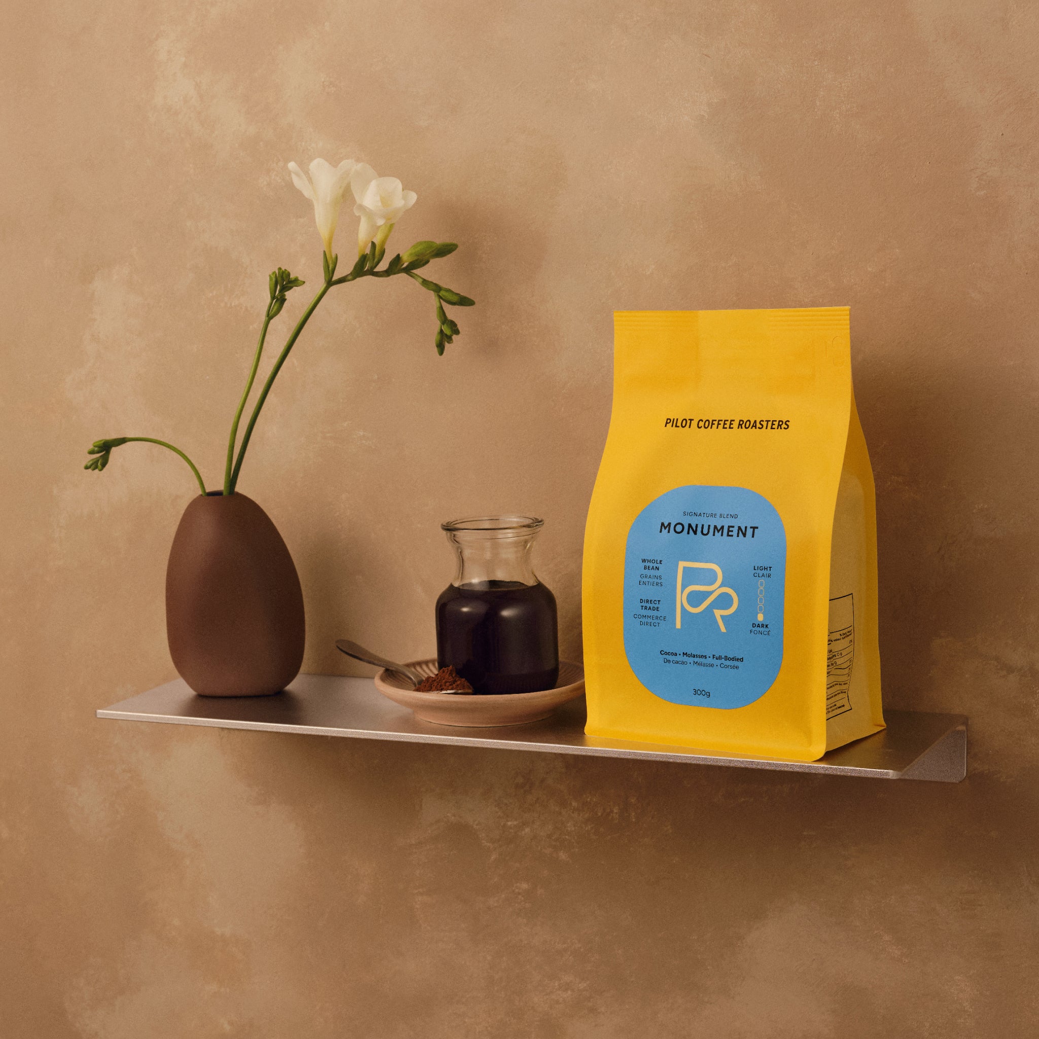 Monument Blend coffee bag with blue label sits beside a vase of white flowers on a shelf against a tan beige background.