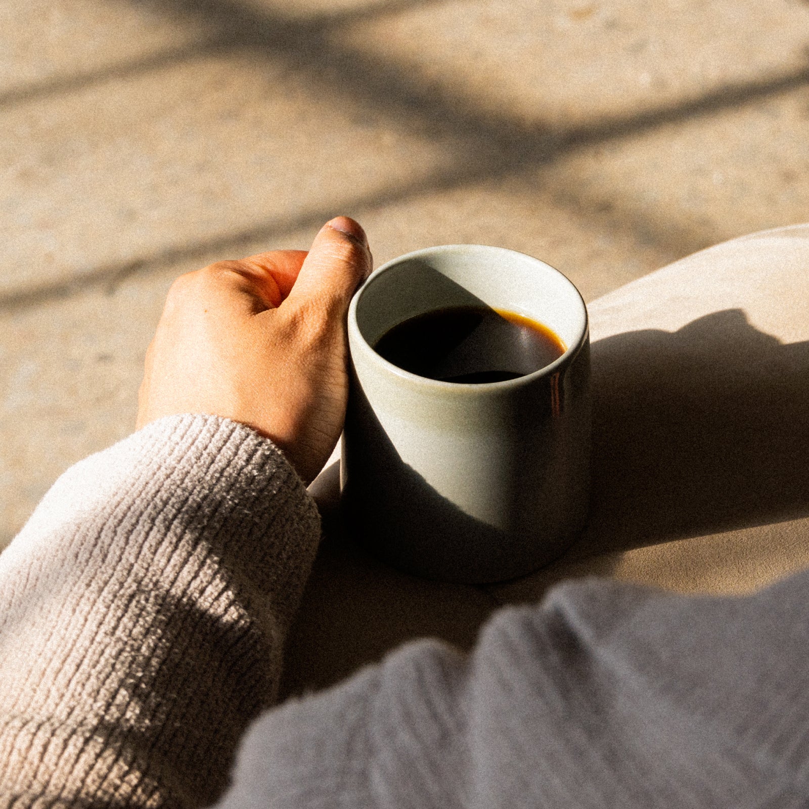 Hand holding a grey mug of black coffee in warm sunlight