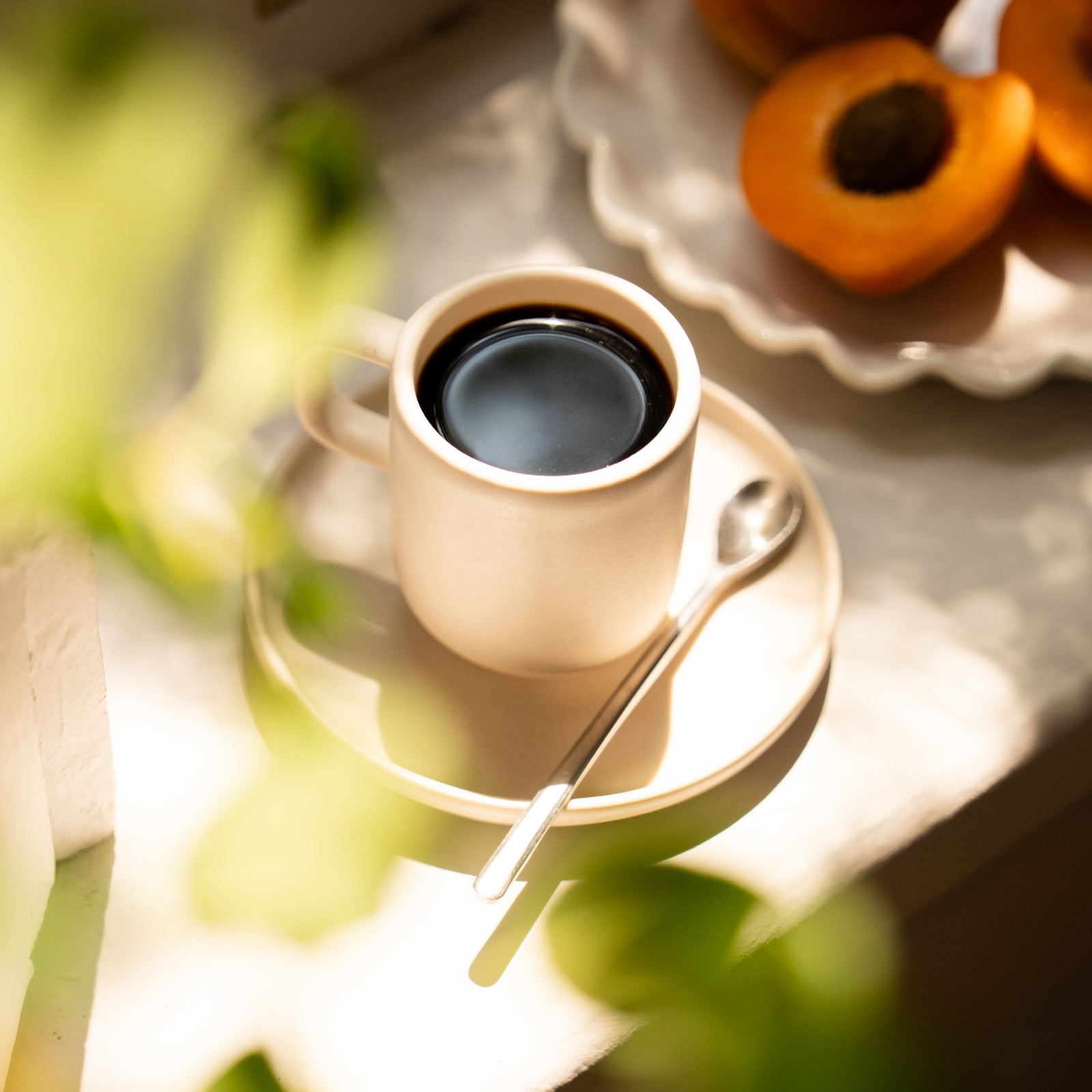 Espresso in a white cup on a saucer with a metal spoon, beside a plate of halved apricots in warm light