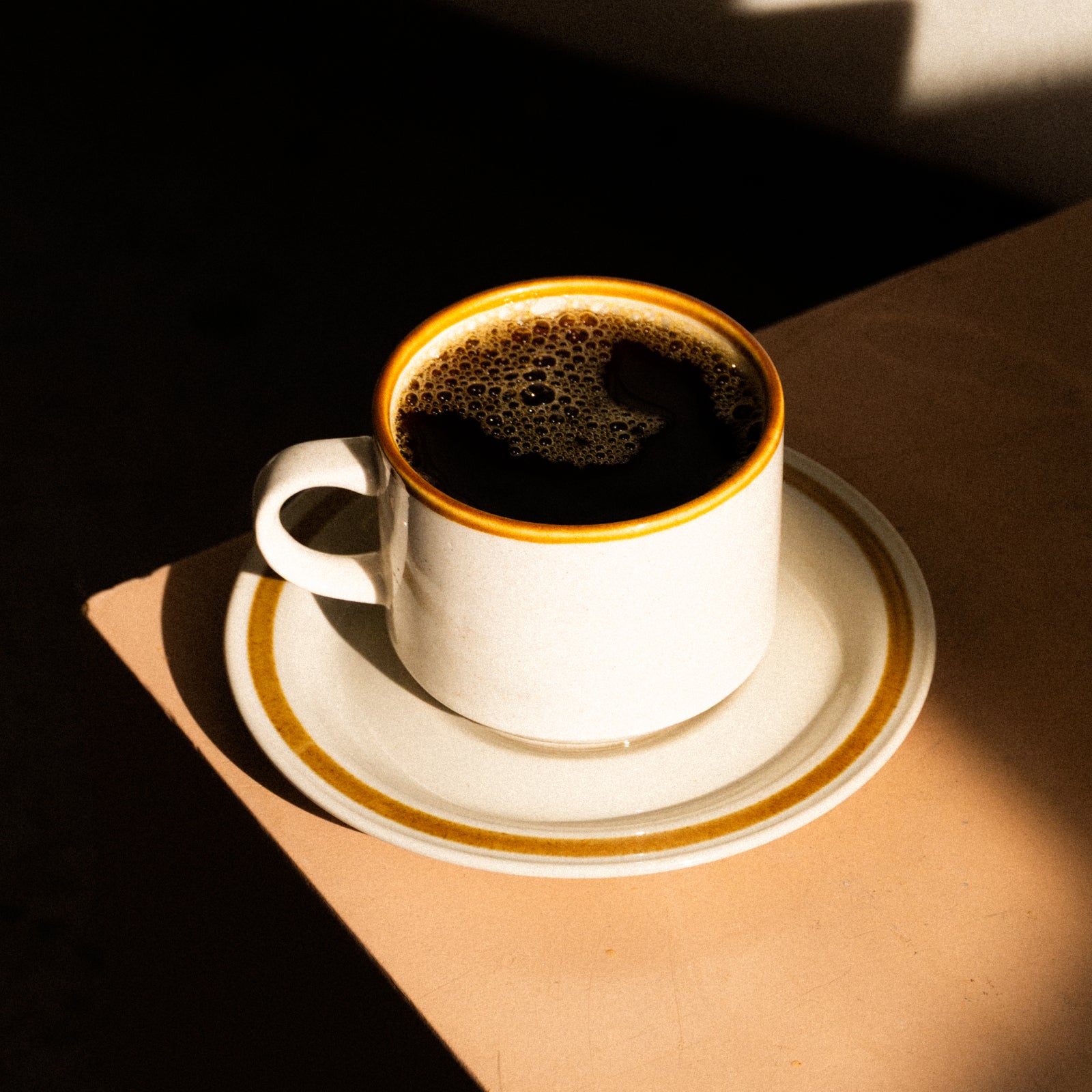 White ceramic coffee cup with a yellow-rimmed edge on a matching saucer, filled with dark coffee, resting on a wood table with warm sunlight