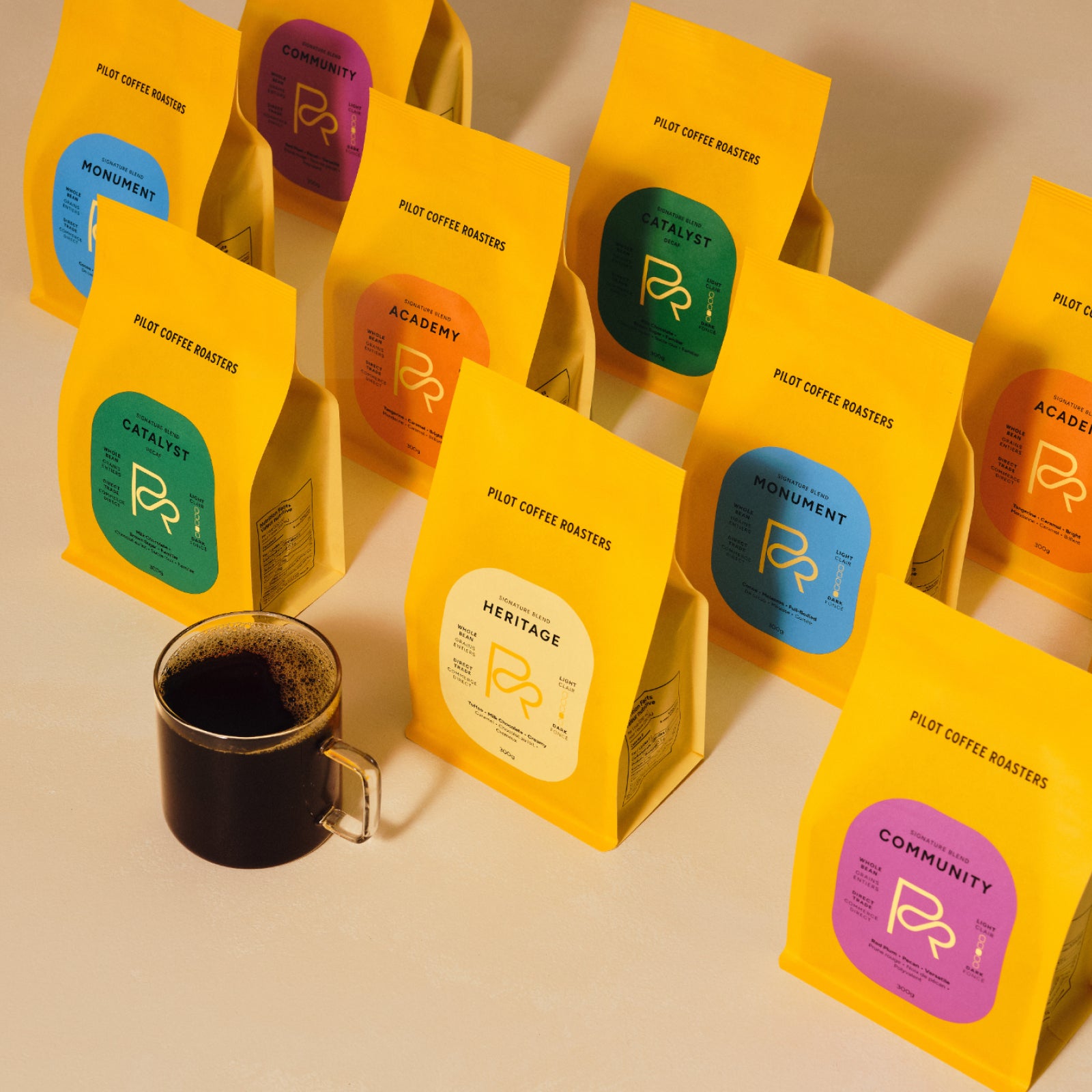 Assortment of bright yellow Pilot Coffee Roasters bags with colorful labels beside a cup of coffee on a table