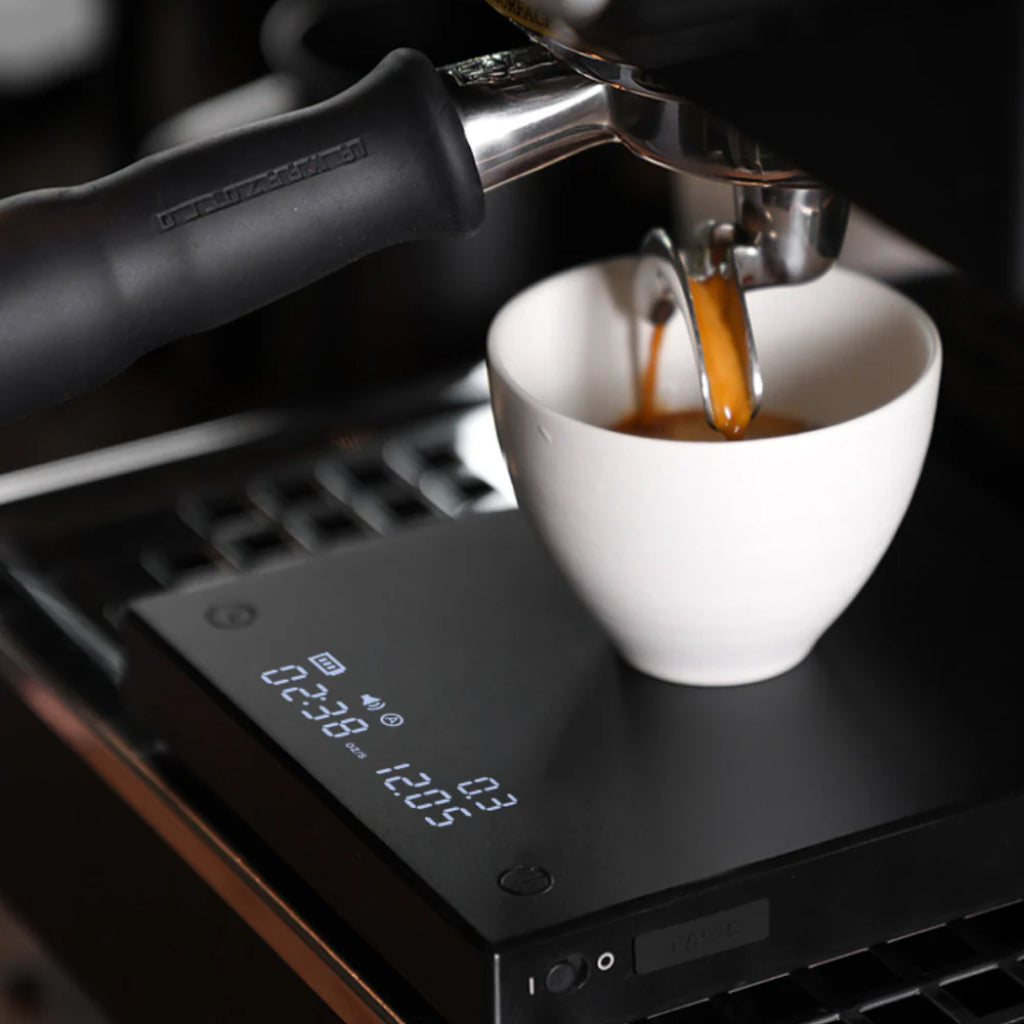 Espresso pours from a portafilter into a white cup on a sleek matte black digital scale with LCD lit display screen.