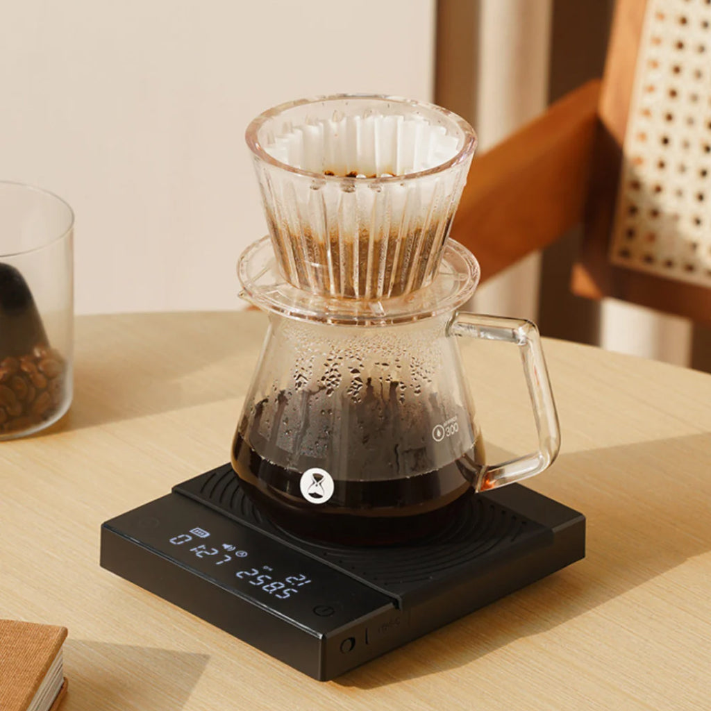 Clear pour over coffee dripper rested on a glass carafe, on a black Timemore Basic 2 digital scale.