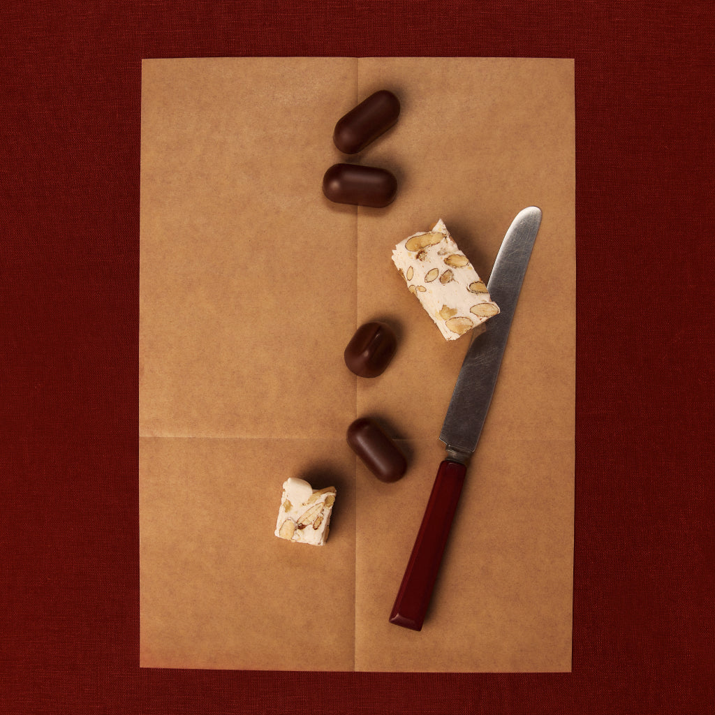 Flat lay of chocolate pieces and an almond nougat bar on brown parchment with a knife.